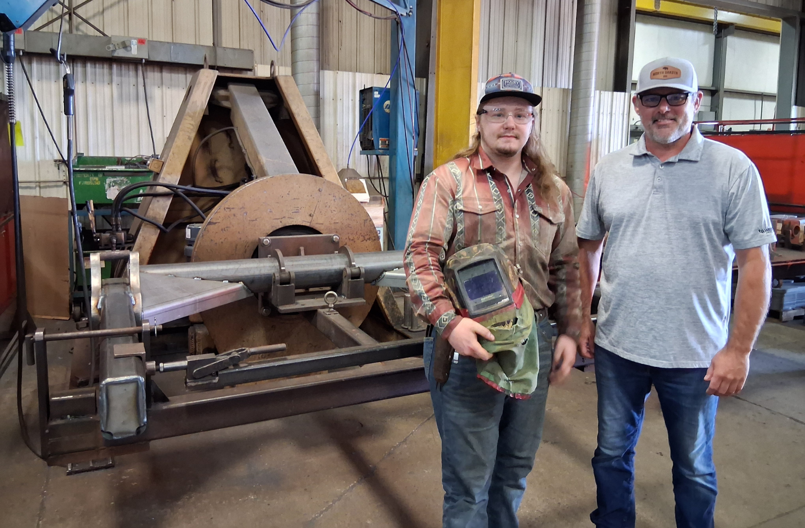 Two men stand near welding machinery in a metal fabrication facility