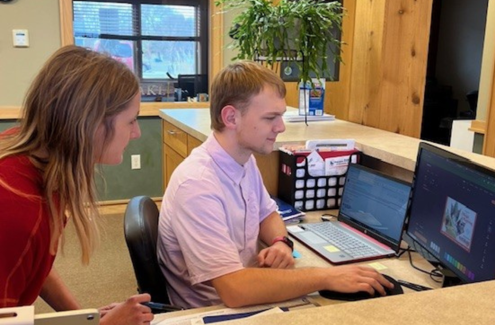A student works on the computer at a business desk while an employee looks on to help him
