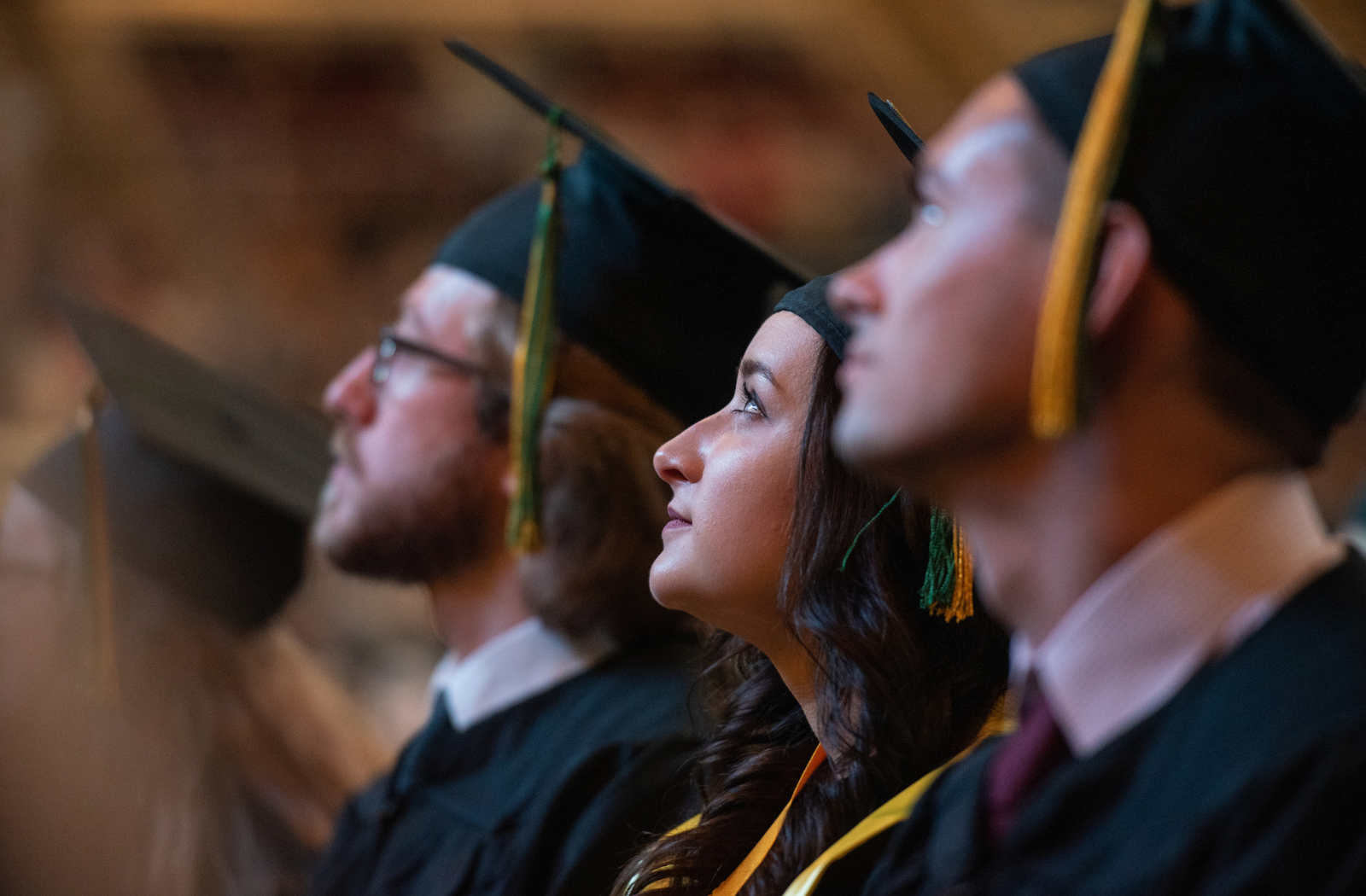 Three students wearing graduation caps and gowns listen to a speaker at graduation