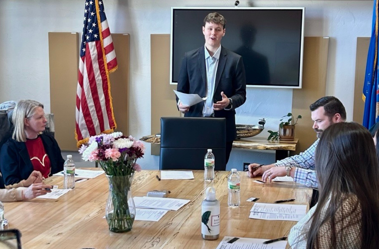A high school student dressed in a suit presents at the front of a table of employees at a meeting