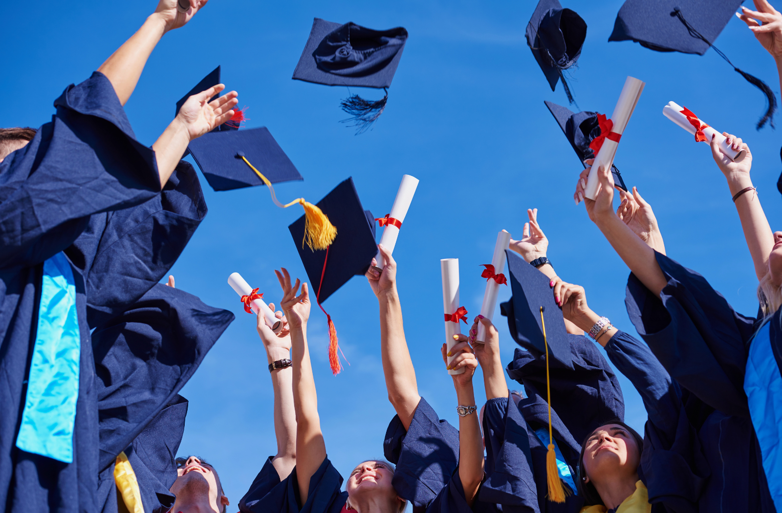 students in graduation robes throw their graduation caps in the air with a blue sky behind them